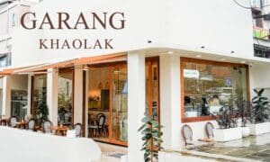 ทัศนียภาพถนนอันมีเสน่ห์ของ Garang Khaolak เผยให้เห็นร้านกาแฟที่แสนอบอุ่น ซึ่งมีหน้าต่างบานใหญ่ กระถางต้นไม้ที่เขียวชอุ่ม และที่นั่งกลางแจ้งที่น่าเชิญชวนที่ตั้งอยู่ในมุมถนนที่มีชีวิตชีวา