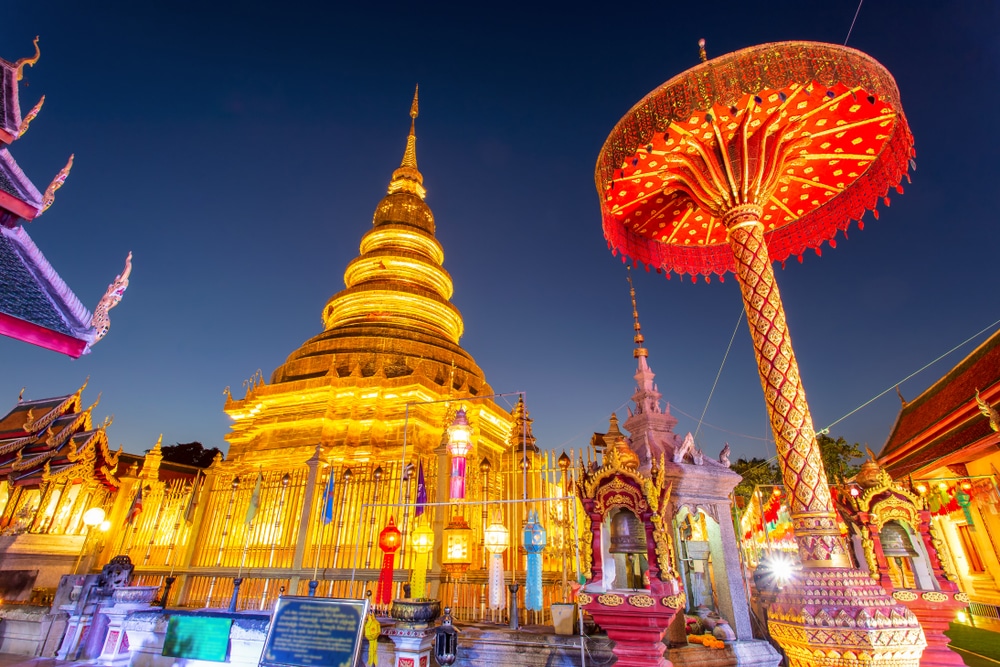 วัดทองคำที่มีรายละเอียดซับซ้อนซึ่งได้รับการส่องแสงในตอนกลางคืน วัดพระธาตุหริภุญชัย โดยมีโครงสร้างอันวิจิตรงดงามและร่มประดับขนาดใหญ่ที่อยู่เบื้องหน้า