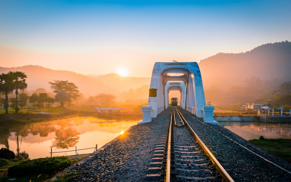 ทางรถไฟที่ทอดผ่านสะพานโค้งสีขาว  ท่องเที่ยวลำพูน พร้อมพระอาทิตย์ขึ้นเหนือเนินเขาและแม่น้ำอันเงียบสงบในยามเช้าที่มีหมอก