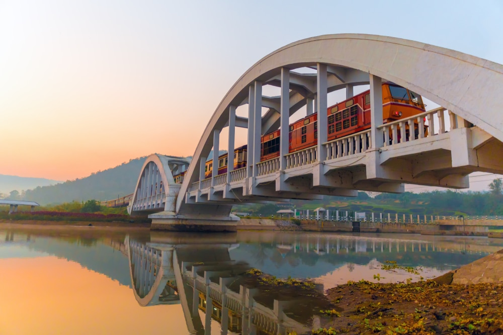 รถไฟวิ่งข้ามสะพานโค้งสีขาวเหนือแม่น้ำอันเงียบสงบขณะพระอาทิตย์ขึ้น โดยมี ท่องเที่ยวลำพูน เนินเขาเป็นฉากหลัง