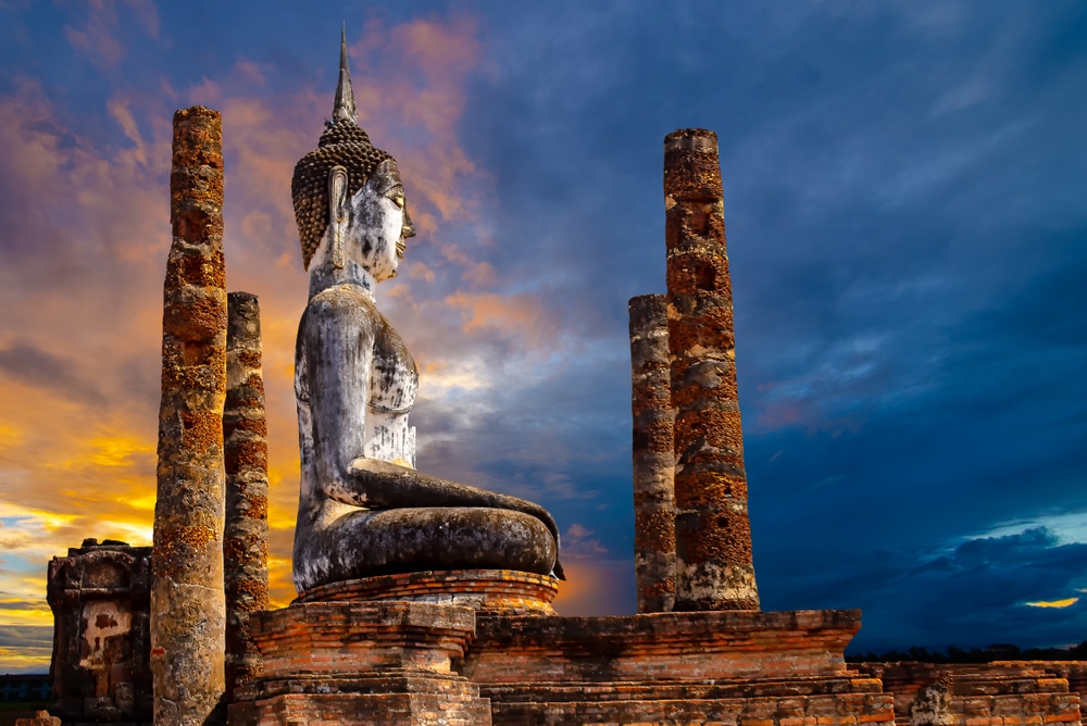 ด้านข้างของพระพุทธรูปปางสมาธิขนาดใหญ่ ณ วัดพิพัฒน์มงคลสุโขทัย ล้อมรอบด้วยเสาผุกร่อนยามพระอาทิตย์ตกดิน โดยมีท้องฟ้าเป็นฉากหลัง วัดพิพัฒน์มงคล