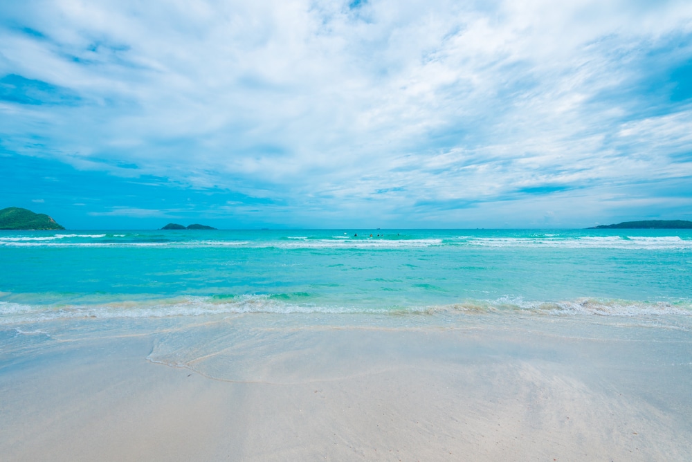 คลื่นสีฟ้าอมเขียวซัดสาดเข้าชายหาดทรายใต้ท้องฟ้าที่มีเมฆบางส่วน มองเห็นเกาะต่างๆ ไกลๆ บนขอบฟ้า ชวนให้นึกถึงการพักผ่อนที่เงียบสงบ ท่องเที่ยวกรุงเทพ ระหว่างสำรวจอัญมณีอันเงียบสงบ เช่น ที่ท่องเที่ยวลำพูน