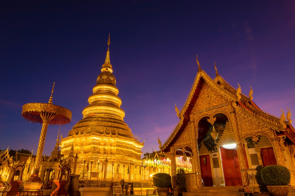 เจดีย์สีทองและอาคารวัดอันวิจิตรงดงามที่ส่องสว่างในช่วงพลบค่ำ วัดพระธาตุหริภุญชัย ตัดกับท้องฟ้าสีม่วงสดใส