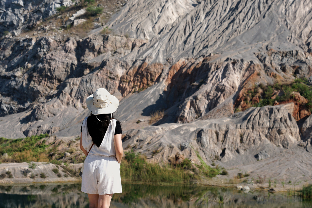 10 สถานที่ท่องเที่ยวราชบุรี คนในชุดขาวและหมวกยืนอยู่ใกล้ ท่องเที่ยวราชบุรี ภูมิประเทศที่เต็มไปด้วยหินและผืนน้ำเล็กๆ จ้องมองหน้าผา จับภาพความงามอันเงียบสงบของพื้นที่ท่องเที่ยวจังหวัดลำพูน