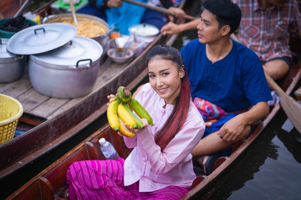 ผู้หญิงคนหนึ่งบนเรือ ท่องเที่ยวจังหวัดราชบุรี ท่องเที่ยวราชบุรี ที่ตลาดน้ำ ล้อมรอบไปด้วยหม้อและผู้คนมากมาย ถือกล้วยหนึ่งกำ ค้นพบฉากที่มีชีวิตชีวาแห่งนี้เป็นส่วนหนึ่งของการเดินทางสำรวจเสน่ห์และวัฒนธรรมเมื่อคุณท่องเที่ยวลำพูน