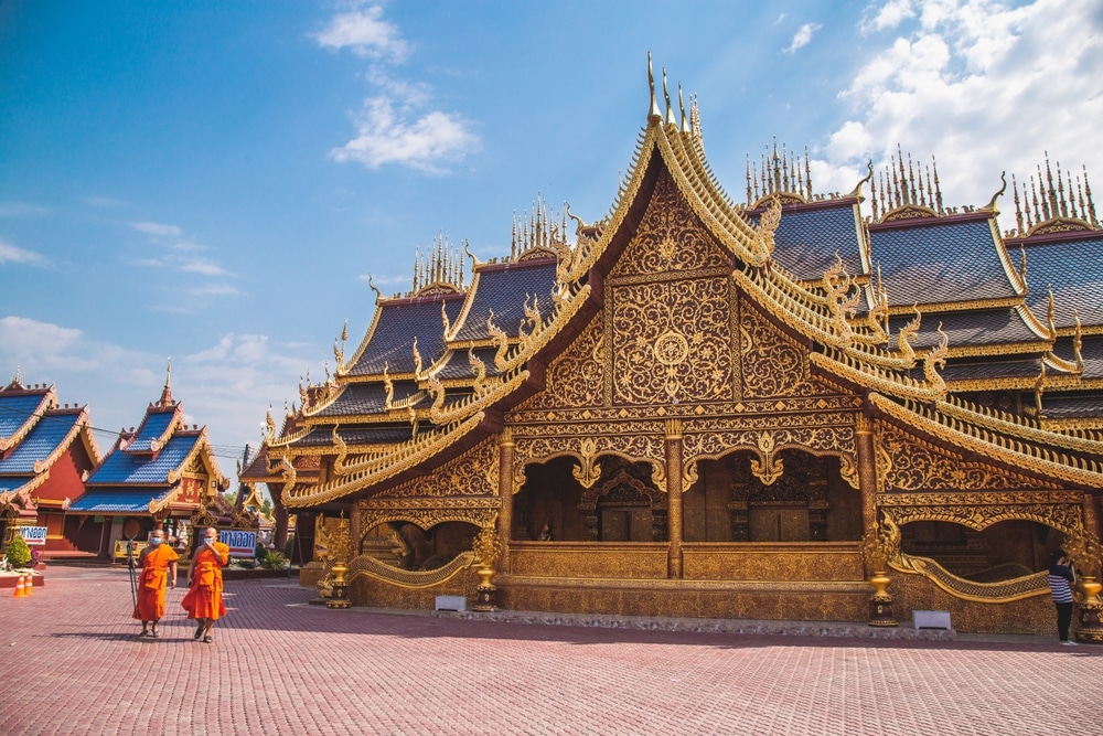 พระสงฆ์ 3 รูปสวมจีวรสีส้มเดินผ่านวัดพิพัฒน์มงคล สีทองอร่าม ประดับด้วยงานแกะสลักอันวิจิตรประณีต วัดพิพัฒน์มงคล และหลังคากระเบื้องสีฟ้า ท่ามกลาง วัดพิพัฒน์มงคล ท้องฟ้าที่มีเมฆบางส่วน