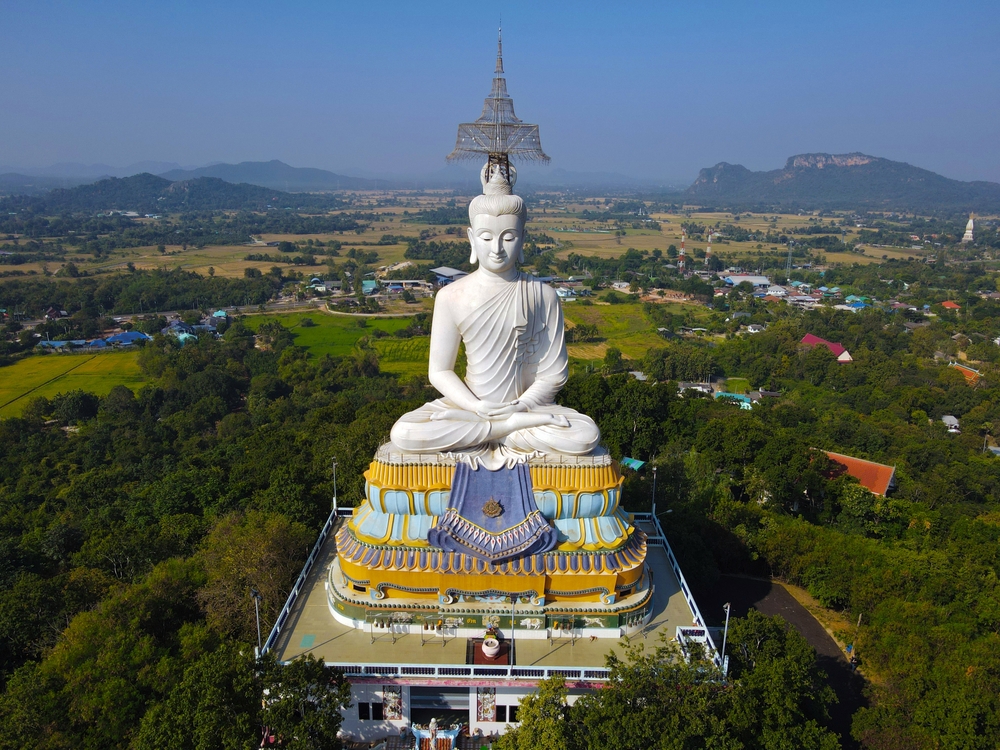 มุมมองทางอากาศของพระพุทธรูปสีขาวขนาดใหญ่นั่งบนแท่นสีสันสดใส  ท่องเที่ยวราชบุรี  ล้อมรอบด้วยแมกไม้เขียวขจีและเนินเขาห่างไกลภายใต้ท้องฟ้าสีฟ้าใส จับภาพความงามอันเงียบสงบของพื้นที่ท่องเที่ยวจังหวัดลำพูน