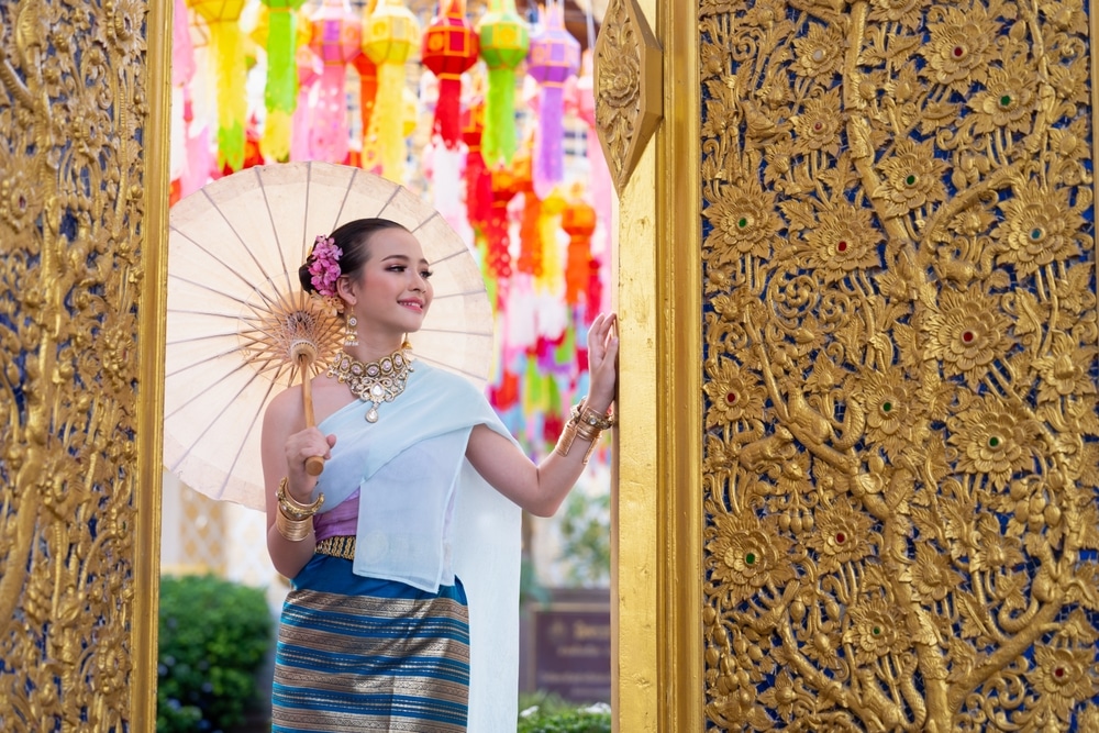 ผู้หญิงสวมชุดประจำชาติ วัดพระธาตุหริภุญชัย ถือร่มยืนอยู่หน้าประตูสีทองประดับตกแต่งซึ่งมีโคมไฟสีสันสดใสอยู่เบื้องหลัง