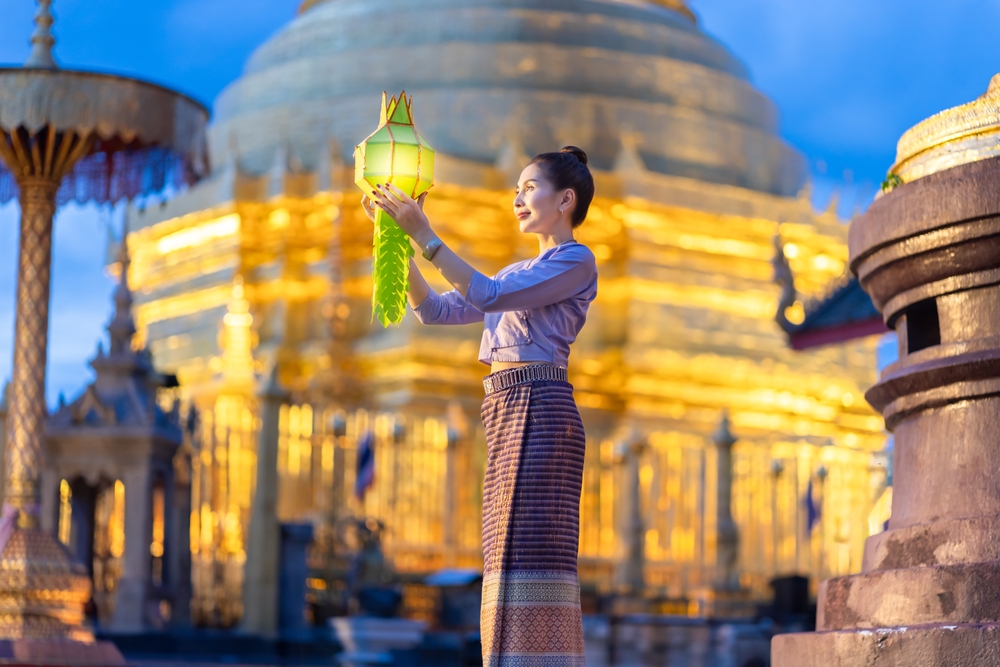 สตรีผู้สวมชุดประจำชาติถือโคมสีเขียว ท่องเที่ยวลำพูน โดยมีฉากหลังเป็นวัดสีทองยามพลบค่ำ