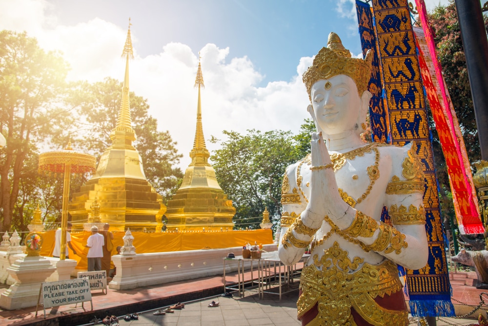 รูปปั้นทองคำในท่าพนมมือตั้งตระหง่านอย่างสง่างามที่ ดอยเชียงราย ด้านหน้าวิหารอันวิจิตรงดงามภายใต้ท้องฟ้าสดใส ใกล้ๆ กันมีป้ายขอให้นักท่องเที่ยวถอดรองเท้า