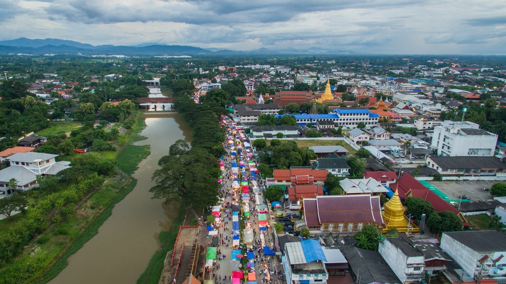 มุมมองทางอากาศของตลาดริมถนนที่คึกคักในเมือง ตลาดแห่งนี้เรียงรายไปด้วยเต็นท์หลากสีสันริมคลอง โดยมี วัดพระธาตุหริภุญชัย อาคารและภูเขารายล้อมอยู่เบื้องหลังภายใต้ท้องฟ้าที่มีเมฆครึ้ม