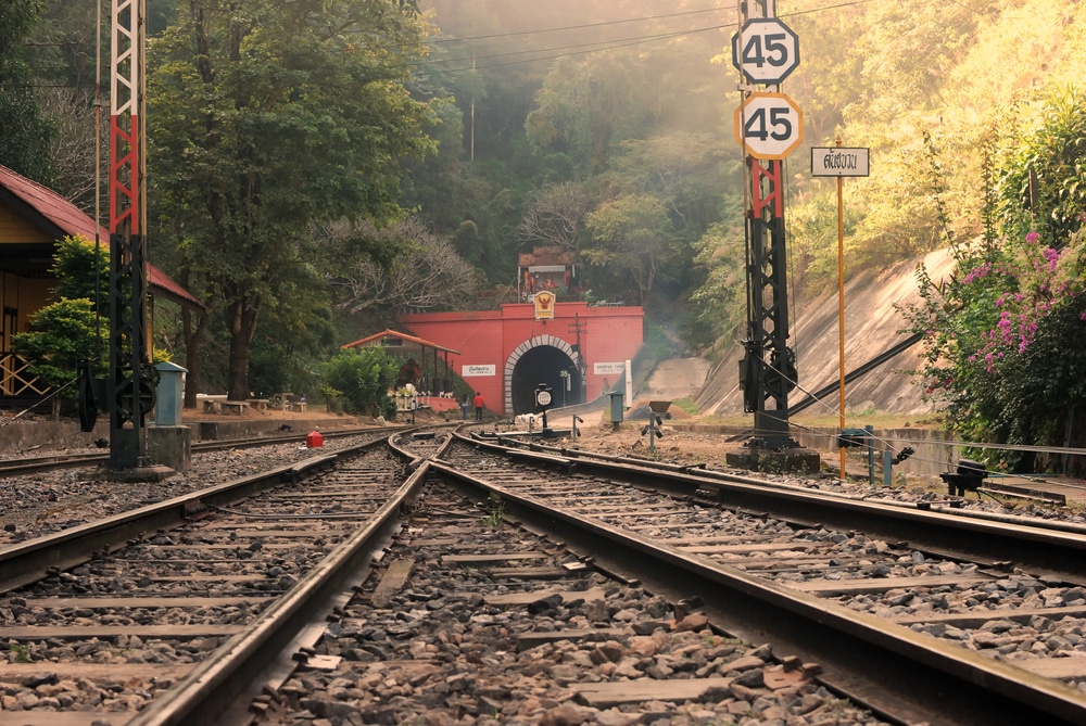 รางรถไฟที่นำไปสู่ทางเข้าอุโมงค์สีแดงที่ล้อมรอบด้วยต้นไม้และใบไม้ มีป้ายจำกัด ท่องเที่ยวลำพูน ความเร็ว 2 ป้ายแสดง 45 สถานที่ท่องเที่ยวจังหวัดลำพูน