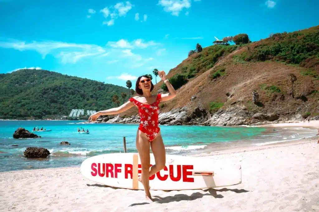 คนในชุดว่ายน้ำสีแดงโพสท่าอย่างสนุกสนานบนหาดทรายข้างกระดาน "Surf Rescue" โดยมีน้ำทะเลสีฟ้าครามและเนินเขาเขียวขจีเป็นฉากหลัง ถ่ายทอดแก่นแท้ของ เที่ยวทะเลภูเก็ต