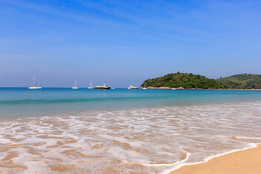 ชายหาดทรายขาว คลื่นเบาๆ น้ำทะเลสีฟ้าใส และเรือหลายลำจอดทอดสมออยู่ ทะเลภูเก็ต ใกล้เกาะสีเขียวเล็กๆ ใต้ท้องฟ้าใส โชว์ความงามอันน่าทึ่งของ ทะเลภูเก็ตอันงดงาม.