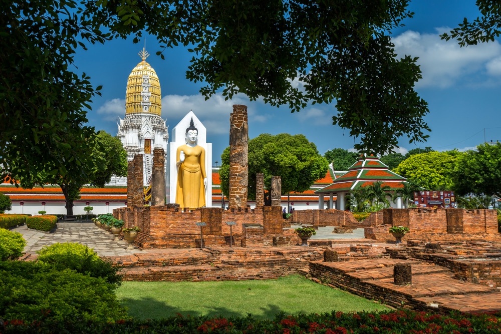 ในบรรยากาศอันเงียบสงบนอกกรุงเทพฯ ซากอิฐโบราณมีพระพุทธรูปยืนคู่บารมีและเจดีย์สีทอง ท่องเที่ยวภาคกลาง เป็นพื้นหลัง ทั้งหมดนี้ล้อมรอบด้วยต้นไม้และสวนอันเขียวชอุ่ม เหมาะสำหรับการท่องเที่ยวหน้าฝนใกล้กรุงเทพผจญภัย