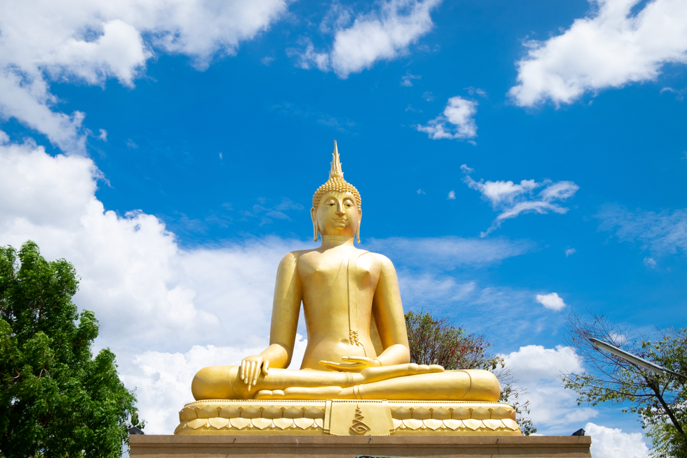 พระพุทธรูปทองคำองค์ใหญ่ประทับอยู่ใต้ท้องฟ้าสีครามสดใสและมีเมฆสีขาวรายล้อมไปด้วยต้นไม้ ณ วัดที่ลพบุรี อันเงียบสงบ