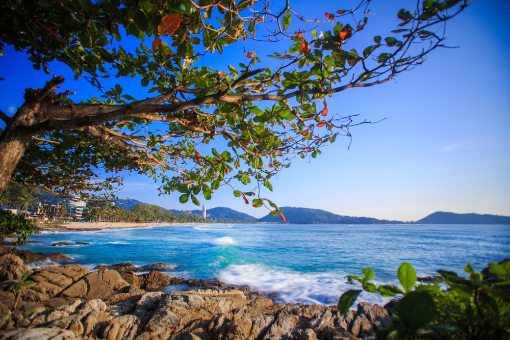 ชายหาดที่สวยงามพร้อมชายฝั่งหิน น้ำทะเลสีฟ้าครามอันเงียบสงบ และต้นไม้ที่มีใบไม้สีเขียวอยู่เบื้องหน้าแสดงถึงเสน่ห์ของการท่องเที่ยวทะเลภูเก็ต เนินเขาอันห่างไกลใต้ท้องฟ้าสีครามช่วยเติมเต็มภูมิทัศน์ทะเลภูเก็ตที่สวยงามแห่งนี้