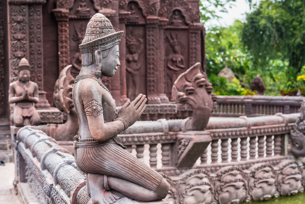 รูปปั้นหินรูปคนคุกเข่าพนมมืออธิษฐาน ตั้งอยู่ในโครงสร้างคล้ายวัด ประดับด้วยงานแกะสลักและองค์ประกอบตกแต่งอันวิจิตรงดงาม ล้อมรอบด้วยต้นไม้เขียวขจี ชวนให้นึกถึงบรรยากาศเงียบสงบที่ วัดลพบุรี