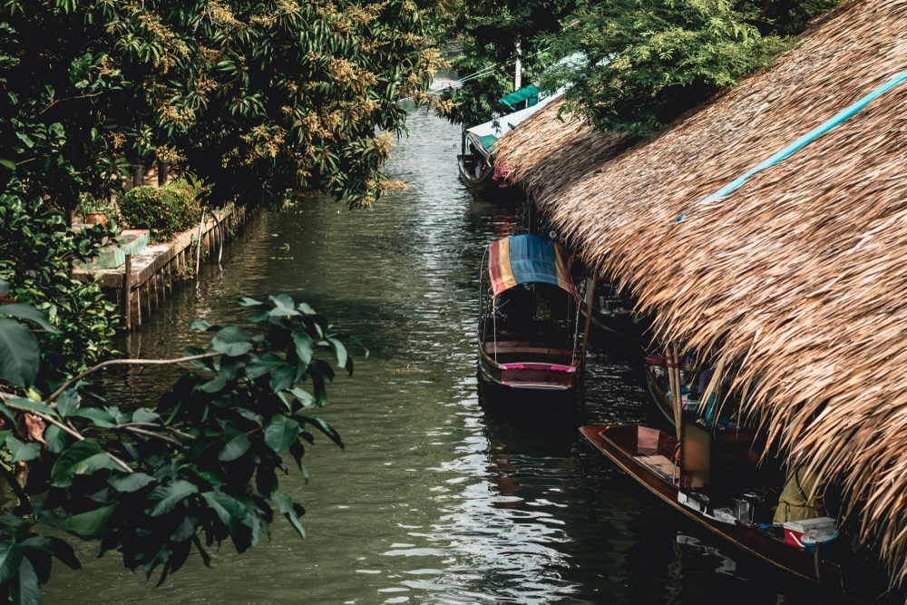 เรือจอดเทียบท่า ตลาดน้ำกรุงเทพ ตามคลองแคบๆ ที่มีความเขียวขจีและมีโครงสร้างหลังคามุงจากทางด้านขวา ชวนให้นึกถึงตลาดน้ำในกรุงเทพซึ่งมีแผงขายของที่มีชีวิตชีวาเรียงรายอยู่ริมทางน้ำ