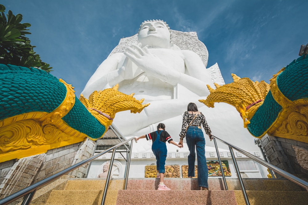 คนสองคนจับมือกันขึ้นบันได วัดสวยลพบุรี ไปยังรูปปั้นสีขาวขนาดใหญ่ของคนนั่งที่วัดดังลพบุรี โดยมีรูปปั้นมังกรสีสันสดใสเรียงรายอยู่ราวบันได