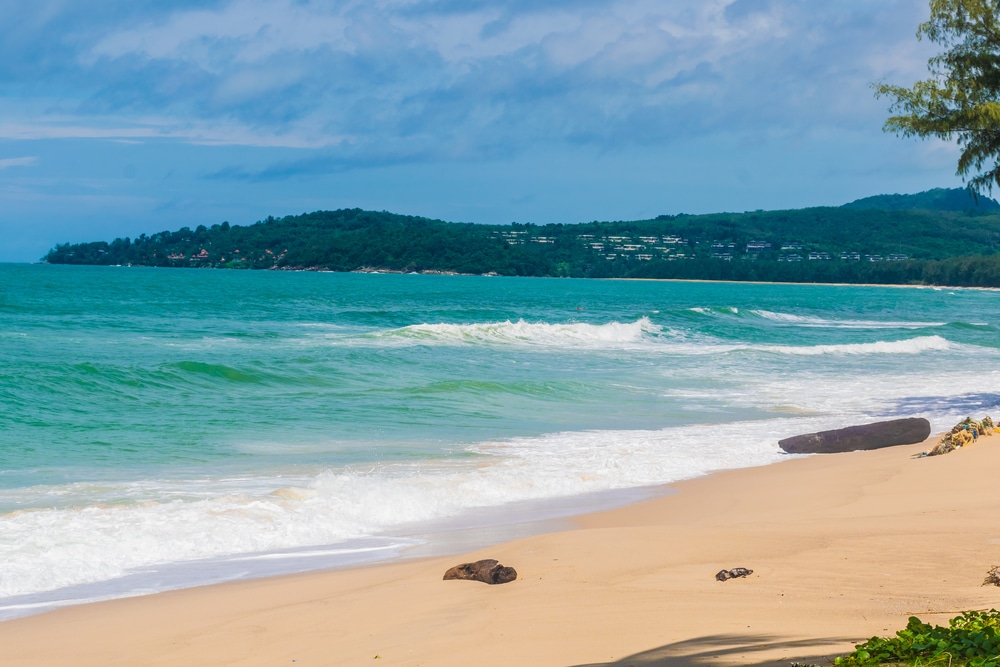 หาดทรายที่มีคลื่นอ่อนโยนและเนินเขาเขียวขจีเป็นพื้น ทะเลภูเก็ต หลังภายใต้ท้องฟ้าที่มีเมฆเป็นบางส่วน เผยให้เห็นความงามของทะเลทะเลภูเก็ตสวยๆ