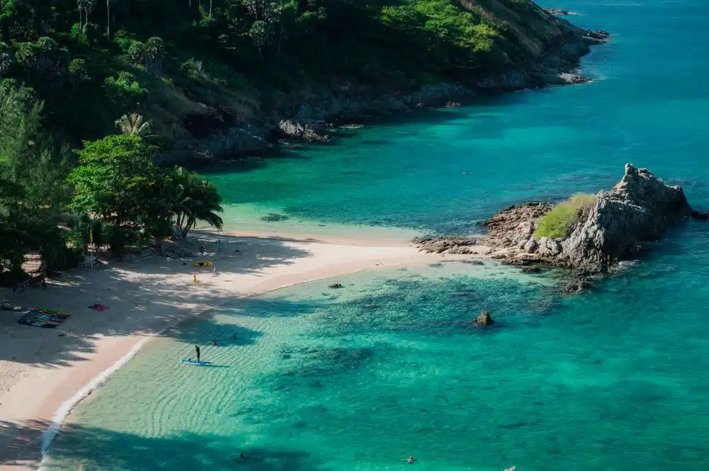 มุมมองทางอากาศของอ่าวสีฟ้าครามใน ทะเลภูเก็ต สวยงาม มีหาดทราย หินโผล่ และต้นไม้เขียวขจี ผู้คนไม่กี่คนกำลังว่ายน้ำ ในขณะที่คนหนึ่งกำลังเล่นซับบอร์ดใกล้ชายฝั่ง