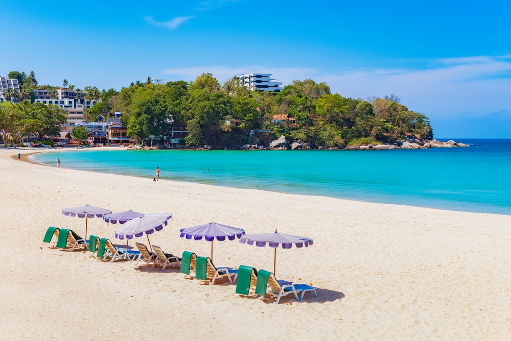 ชายหาดทรายสีฟ้าครามมีเก้าอี้ชายหาดหลายตัวใต้ร่มสีม่วง ต้นไม้และอาคารต่างๆ มองเห็นได้เป็นฉากหลังภายใต้ท้องฟ้าสีฟ้าใส เป็นฉากที่สมบูรณ์แบบสำหรับผู้ที่ใฝ่ฝันอยากเที่ยวทะเลภูเก็ต