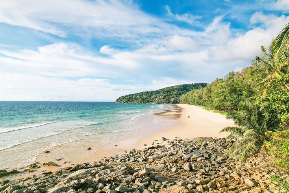 ชายหาดที่มีแสงแดดสดใสในเที่ยวทะเลภูเก็ตที่มีหาดทรายสีทอง แนวชายฝั่งหิน และต้นไม้เขียวชอุ่มใต้ ทะเลภูเก็ต ท้องฟ้าสีคราม สองคนเดินชิลๆ ริมน้ำ ชมความงามอันเงียบสงบของทะเลที่ภูเก็ต