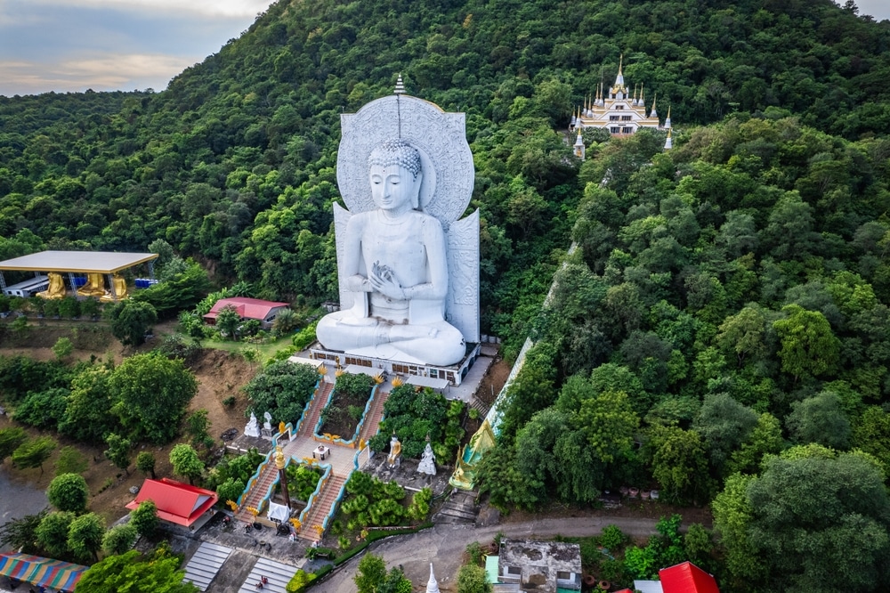 มุมมองทางอากาศของพระพุทธรูป วัดสวยลพบุรี องค์ใหญ่ประทับนั่งบนเนินเขาที่รายล้อมไปด้วยต้นไม้เขียวขจี โดยมีวัดดังลพบุรีอันเลื่องชื่อปรากฏเป็นฉากหลัง