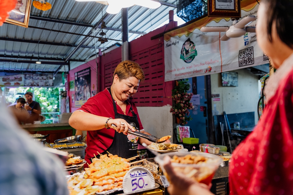 ที่แผงขาย ตลาดน้ำกรุงเทพ ของสุดคึกคักในตลาดน้ำใกล้กรุงเทพ พ่อค้าเสื้อแดงเสิร์ฟอาหารหลากหลายเมนู ป้ายที่แสดง "50" ดึงดูดสายตาท่ามกลางฉากที่มีชีวิตชีวา