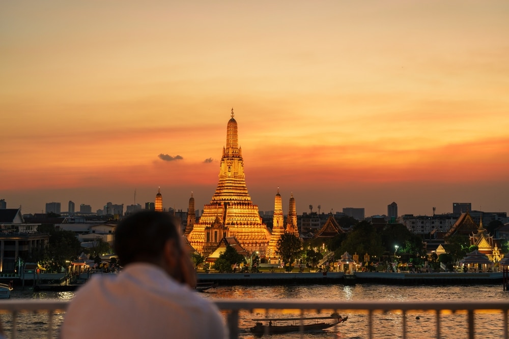 มีคนจ้องมองวัดวัดอรุณที่ส่องสว่างในกรุงเทพฯ ประเทศไทย ยาม สถานที่ท่องเที่ยวภาคกลาง 10 อันดับ พระอาทิตย์ตกดิน ฉากอันน่าทึ่งนี้แสดงให้เห็นแม่น้ำที่อยู่เบื้องหน้าและเส้นขอบฟ้าของเมืองที่อยู่ไกลออกไป เป็นภาพที่น่าจดจำซึ่งเหมาะสำหรับการผจญภัยหน้าฝนใกล้กรุงเทพ