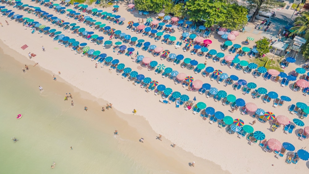 มุมมองทางอากาศของชายหาด ทะเลภูเก็ต ที่พลุกพล่านในทะเลภูเก็ตพร้อมร่มหลากสีสันและผู้คนกำลังผ่อนคลาย คลื่นทะเลซัดหาดทรายเบา ๆ ชวนทุกคนมาสำรวจความสวยงามของเที่ยวทะเลภูเก็ต