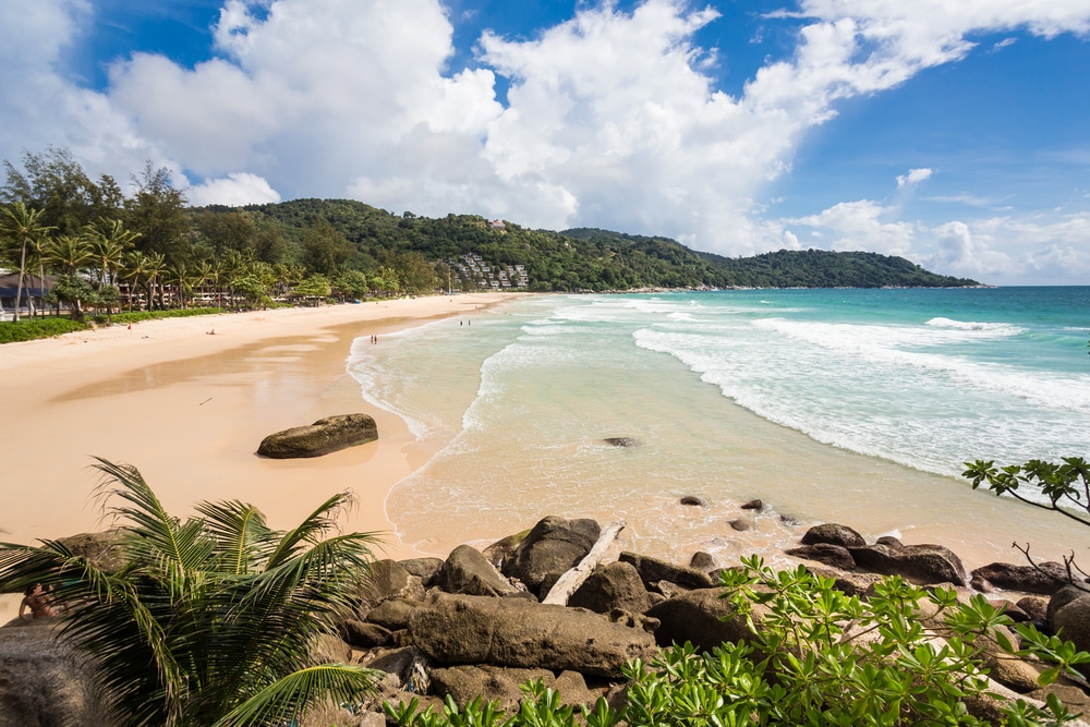 หาดทรายที่มีคลื่นสีฟ้าคราม หินขนาดใหญ่ และเนินเขาสีเขียวชอุ่มภายใต้ท้องฟ้าที่มีเมฆบางส่วนรวบรวมแก่นแท้ของการท่อง เที่ยวทะเลภูเก็ต