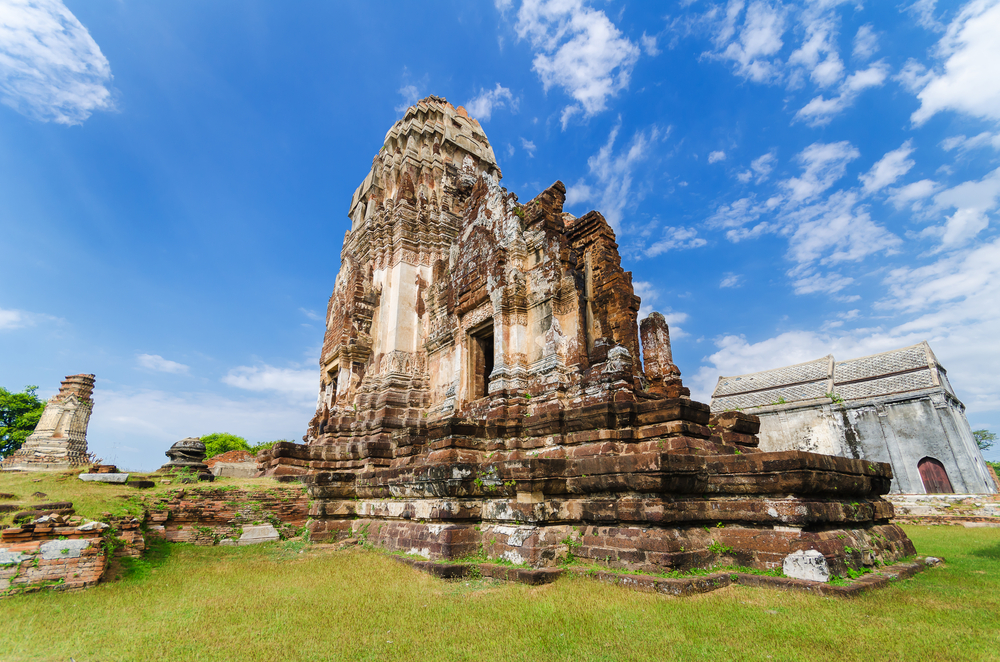 ซากวัดหินโบราณที่วัดลพบุรี ตั้งตระหง่านอยู่ภายใต้ท้องฟ้าสีฟ้าใสที่มีเมฆ วัดสวยลพบุรี กระจัดกระจาย ล้อมรอบด้วยหญ้าเขียวชอุ่มและโครงสร้างขนาดเล็ก