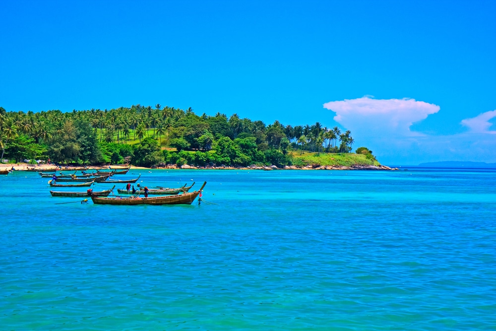 เรือลอยอยู่บนน้ำทะเลสีฟ้าใสของเกาะภูเก็ต ใกล้กับเกาะที่มีต้นไม้เขียวขจีปกคลุมอยู่ท่ามกลางท้องฟ้าสีฟ้าสดใส ทะเลภูเก็ตสวยๆ พร้อมเมฆก้อนใหญ่เพียงก้อนเดียว