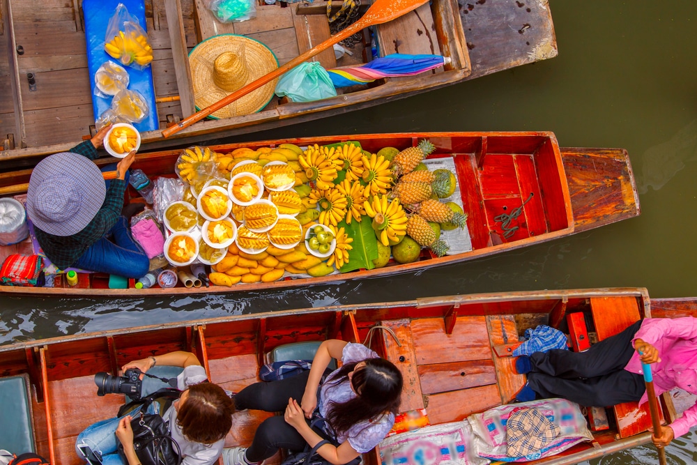 ที่ตลาดน้ำอันคึกคัก ท่องเที่ยวภาคกลาง ใกล้กรุงเทพฯ พ่อค้าแม่ค้าจะนำกล้วย สับปะรด และมะม่วงจากเรือมาขาย แม้ในช่วงหน้าฝนนักท่องเที่ยวจะสำรวจสถานที่อันคึกคักและเก็บภาพบรรยากาศที่มีชีวิตชีวา เหมาะสำหรับการท่องเที่ยวหน้าฝนใกล้กรุงเทพผจญภัย!