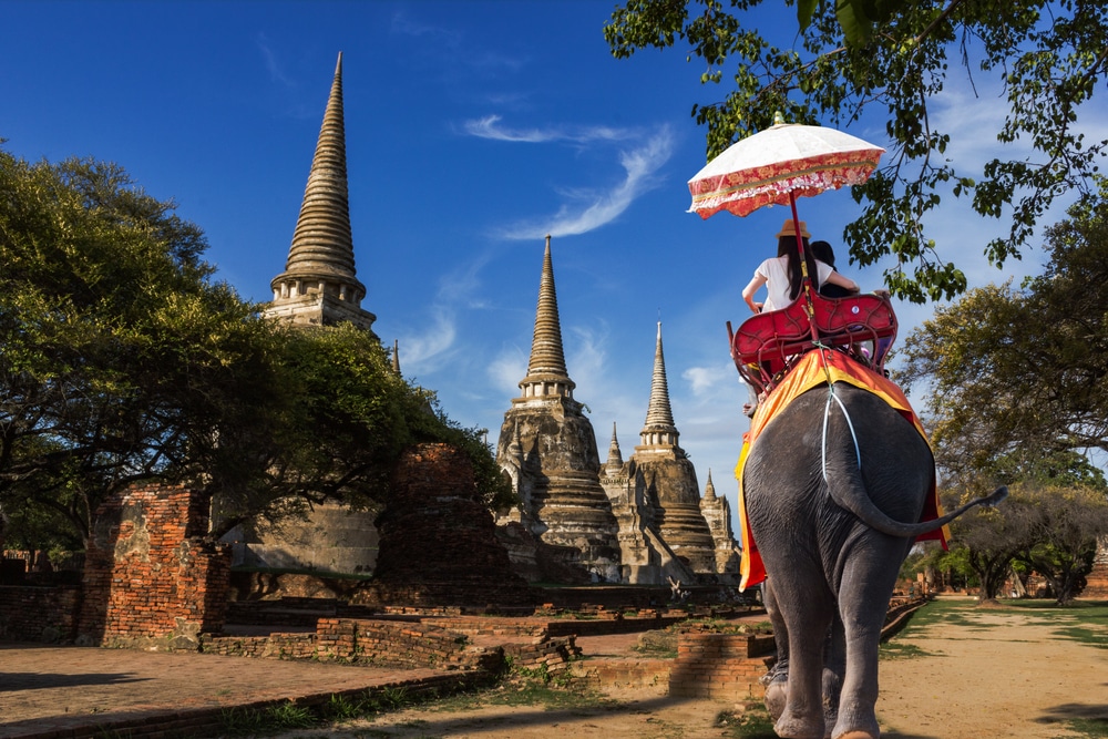 บุคคลหนึ่งขี่ช้าง มรดกโลกในไทย โดยมีร่มกันแดดอยู่หน้าซากปรักหักพังของวัดโบราณ มียอดแหลมแหลมอยู่ใต้ท้องฟ้าสีคราม ฉากอันเงียบสงบนี้เป็นส่วนหนึ่งของมรดกในไทย ซึ่งจัดแสดงมรดกอันยาวนานและความมหัศจรรย์ที่ยูเนสโกไทยปกป้องและเฉลิมฉลอง