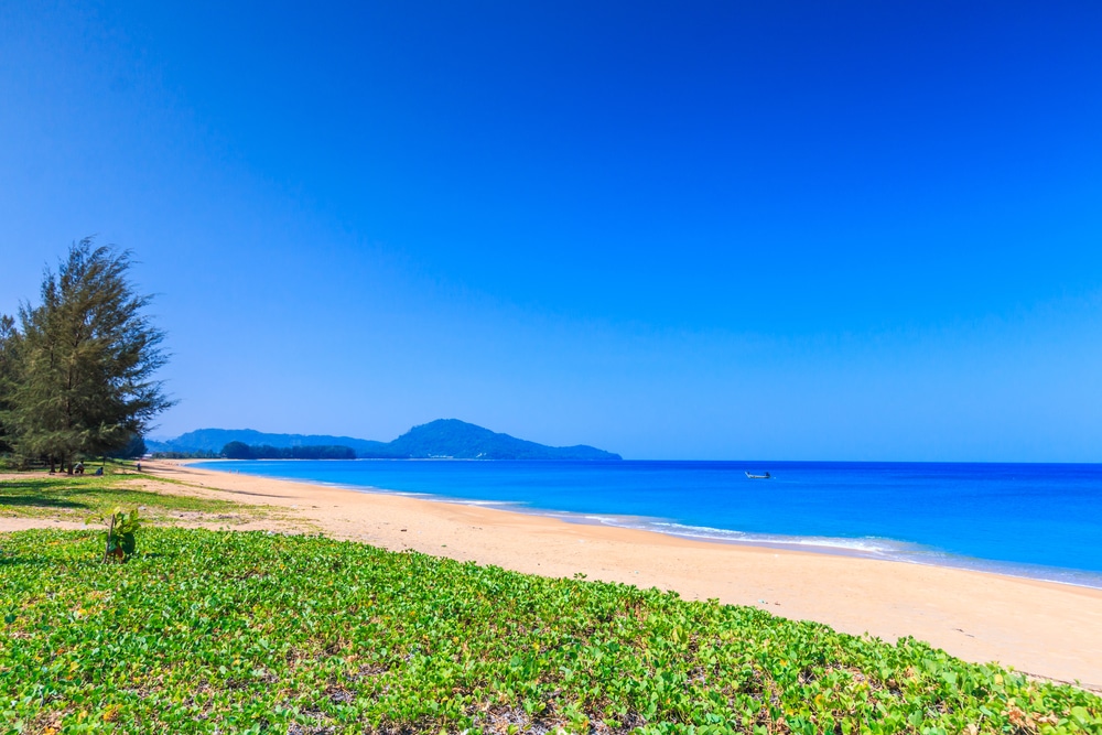 ชายหาดอันเงียบสงบใน ทะเลภูเก็ต ภูเก็ตที่มีทรายสีทอง น้ำทะเลสีฟ้าใส และท้องฟ้าที่สดใส มองเห็นเกาะที่อยู่ไกลออกไปและเรือลำเล็ก ขณะที่ต้นไม้เขียวขจีอยู่ริมชายทะเลอันสวยงามนี้