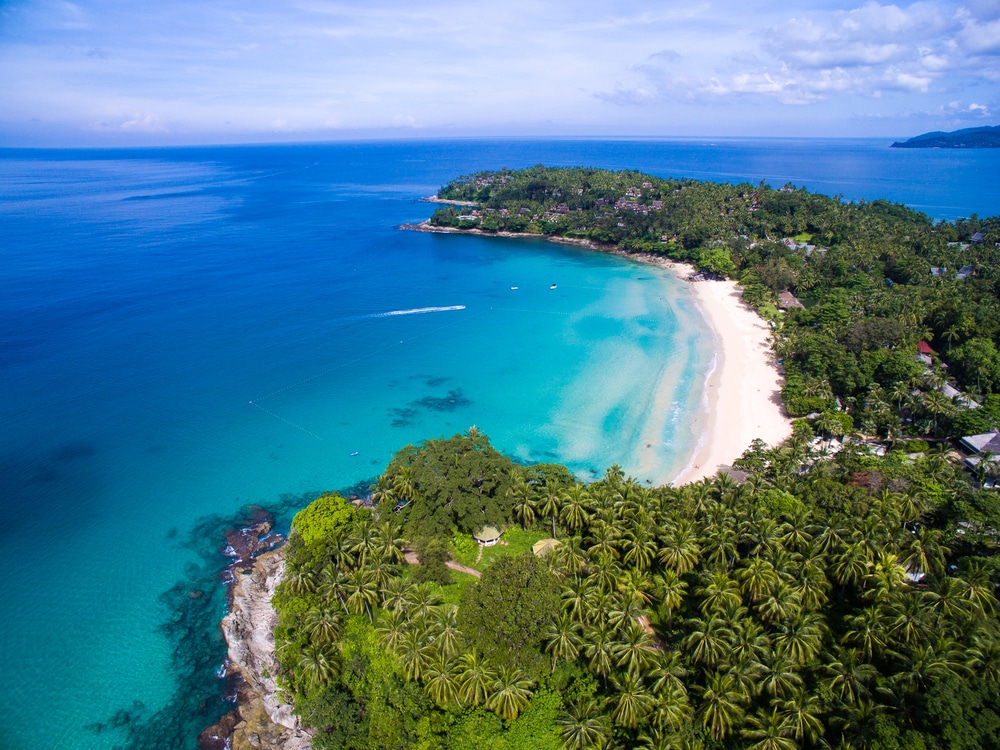 มุมมองทางอากาศของ ทะเลภูเก็ต ชายหาดเขตร้อนในจังหวัดภูเก็ต แสดงให้เห็นน้ำทะเลสีฟ้าใส ต้นไม้เขียวชอุ่ม และแนวชายฝั่งโค้งภายใต้ท้องฟ้าสีฟ้ากว้างใหญ่