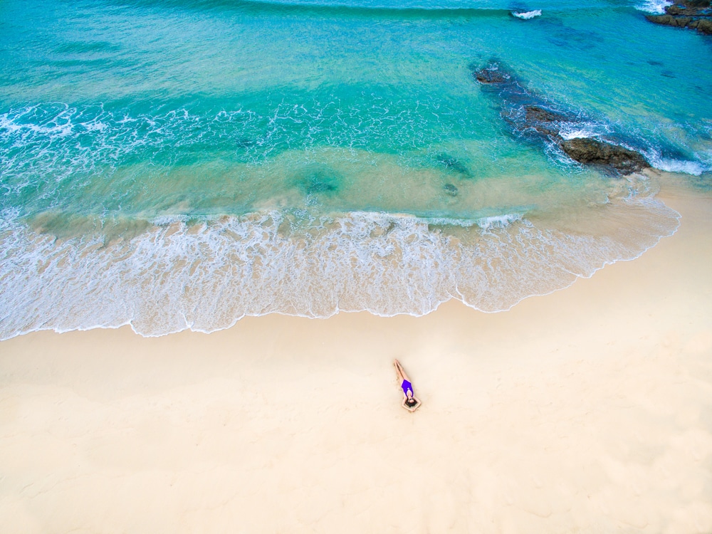 คนในชุดว่ายน้ำนอนเล่นอยู่บน ทะเลภูเก็ต หาดทรายถัดจากคลื่นทะเลสีฟ้าคราม มีก้อนหินให้เห็นในน้ำ ชื่นชมความงามที่น่าทึ่งของ ทะเลภูเก็ตสวยๆ