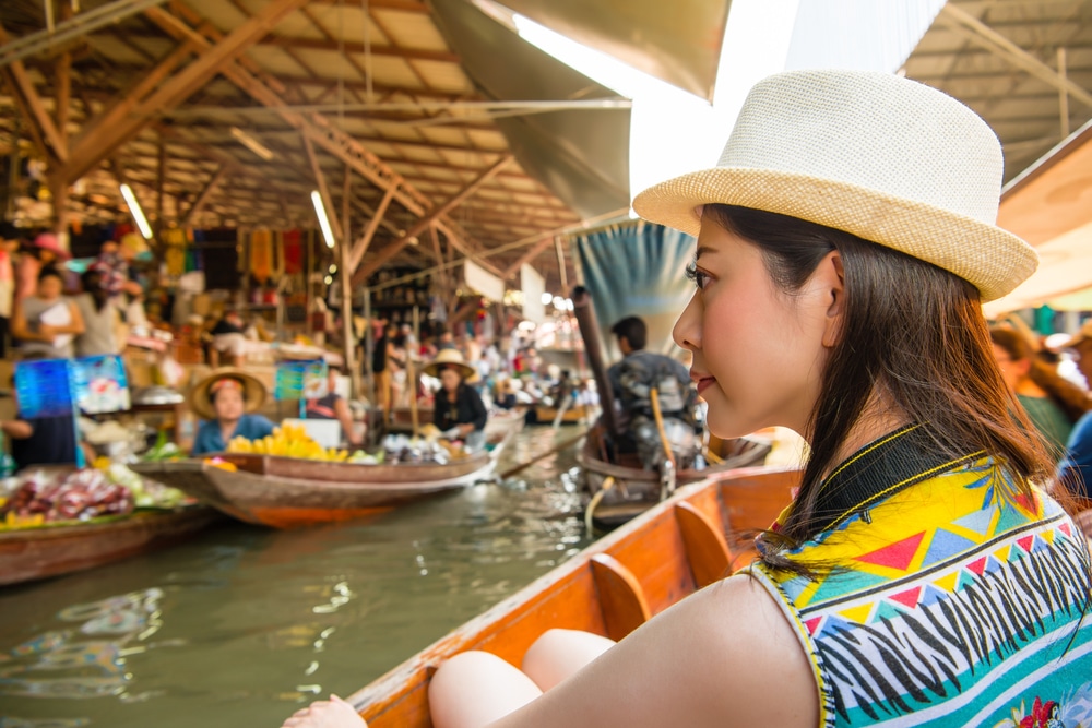 ผู้หญิงสวมหมวกนั่งเรือ ตลาดน้ำกรุงเทพ ที่ตลาดน้ำกรุงเทพ รายล้อมไปด้วยเรือลำอื่นที่เต็มไปด้วยสินค้าและผู้คนพลุกพล่านภายใต้พื้นที่ปิด