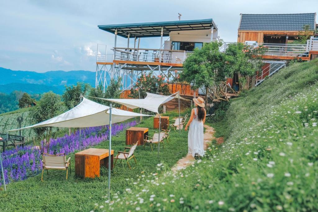 หญิงสาวในชุดสีขาวเดินเล่นไปตามเส้นทางสู่ร้านกาแฟสุดน่ารักบนเนิน ที่พักม่อนแจ่ม เขาที่มีที่นั่งกลางแจ้ง รายล้อมไปด้วยต้นไม้สีเขียวและดอกไม้สีม่วง มองเห็นทิวเขาไกลๆ ใกล้ที่พักม่อนแจ่ม.