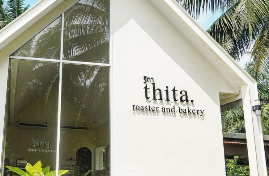 อาคารสีขาวที่มีหลังคาสามเหลี่ยมมีป้าย "th i t a. Roaster and Bakery" ประดับอยู่อย่างภาคภูมิใจ ขณะที่ต้นปาล์มสะท้อนอย่างสง่างามบนกระจกหน้าต่างบานใหญ่.