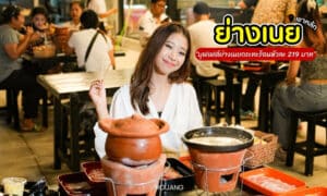 ผู้หญิงกำลังยิ้มอยู่ที่โต๊ะพร้อมอาหารและหม้อดินเผา ภาพร้านอาหารปรากฏให้เห็นในพื้นหลัง ข้อความในภาพเป็นภาษาไทย.