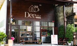 ภายนอกของ Dee Cafe & Eatery คาเฟ่ย่านตลาดเก่า พังงา โดดเด่นด้วยส่วนหน้าอาคารไม้ที่มีเสน่ห์ หน้าต่างกระจกบานใหญ่ กระถางต้นไม้ และป้ายต้อนรับที่เปิดอยู่.