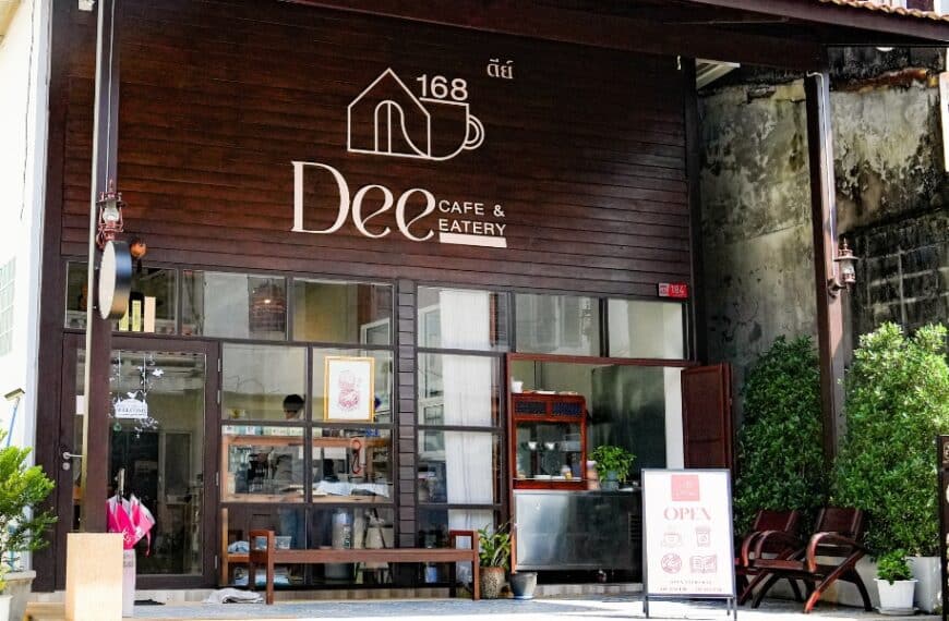 ภายนอกของ Dee Cafe & Eatery คาเฟ่ย่านตลาดเก่า พังงา โดดเด่นด้วยส่วนหน้าอาคารไม้ที่มีเสน่ห์ หน้าต่างกระจกบานใหญ่ กระถางต้นไม้ และป้ายต้อนรับที่เปิดอยู่.