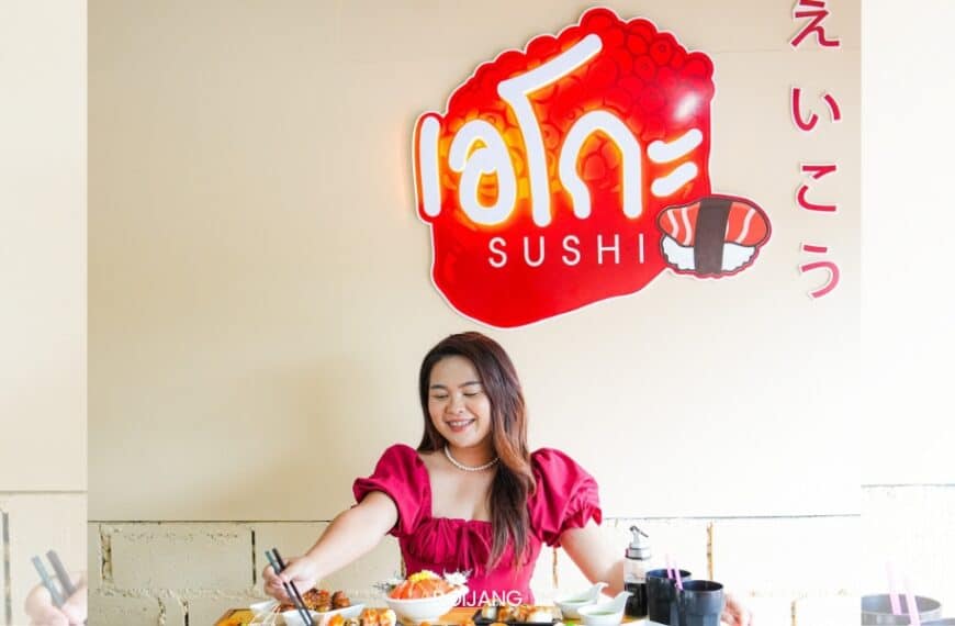 คนใส่เสื้อแดงกำลังนั่งที่โต๊ะที่มีอาหารหลากหลายชนิดในร้าน Eiko Sushi ซึ่งเป็นร้านซูชิสุดเก๋ในตะกั่วป่า ป้ายด้านบนเขียนว่า "ซูชิ" ซึ่งมีการออกแบบที่เก๋ไก๋