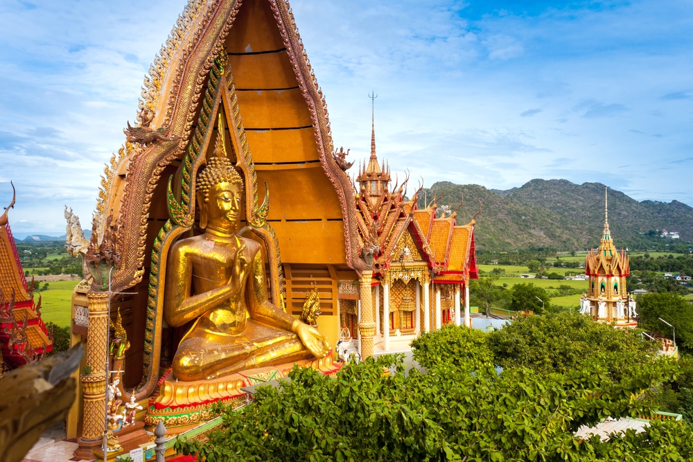 พระพุทธรูปสีทองตั้งตระหง่านอยู่หน้า วัดที่สวยที่สุดในประเทศไทย วัดพระนอนอันวิจิตรงดงาม ตั้งอยู่ท่ามกลางต้นไม้สีเขียวชอุ่มโดยมีภูเขาเป็นฉากหลังภายใต้ท้องฟ้าสีฟ้าใส เชิญชวนผู้มาเยี่ยมชมไหว้พระนอนในบรรยากาศอันเงียบสงบ.