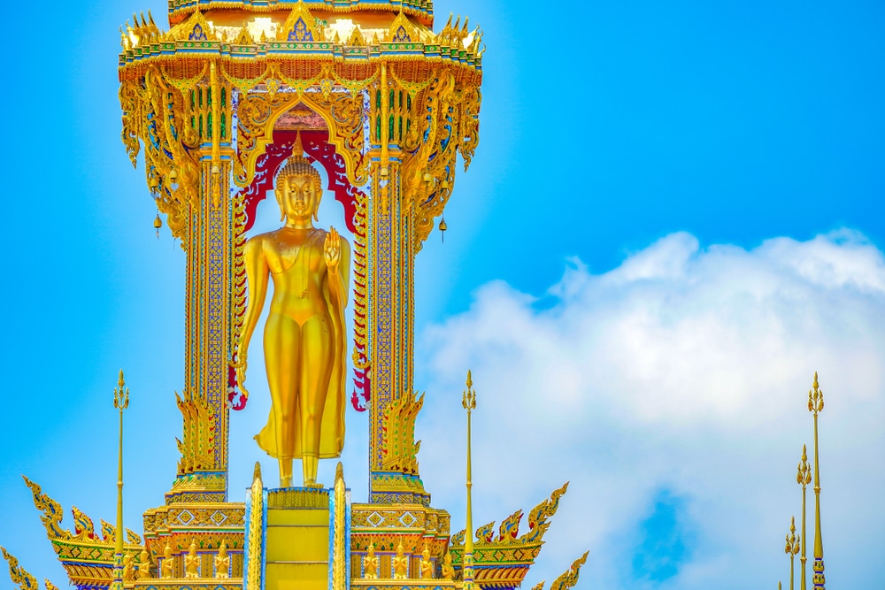  วัดที่สวยที่สุดในประเทศไทย พระพุทธรูปสีทองตั้งตระหง่านอยู่ใต้โครงสร้างหลากสีสันอันวิจิตรงดงาม โดยมีท้องฟ้าสีฟ้าและเมฆสีขาว ชวนให้นึกถึงความเงียบสงบที่วัดพระนอน.