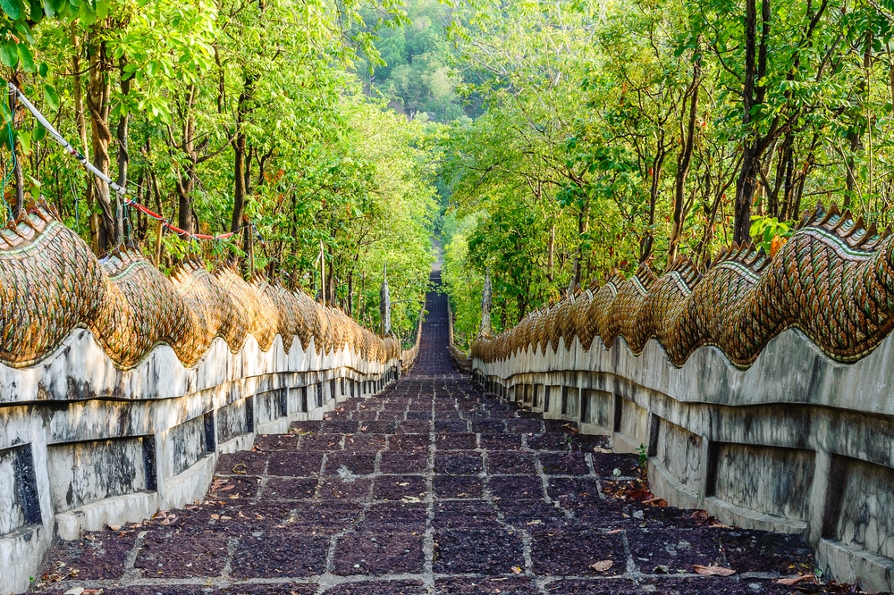 บันไดยาวล้อมรอบ รอยพระพุทธบาท ด้วยรูปปั้นงูอันหรูหราทอดยาวผ่านต้นไม้สีเขียวชอุ่ม นำไปสู่สถานที่ศักดิ์สิทธิ์ของรอยพระพุทธบาท